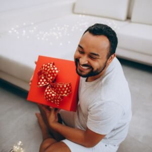Smiling man with a beard holding a red Christmas present with a bow indoors.