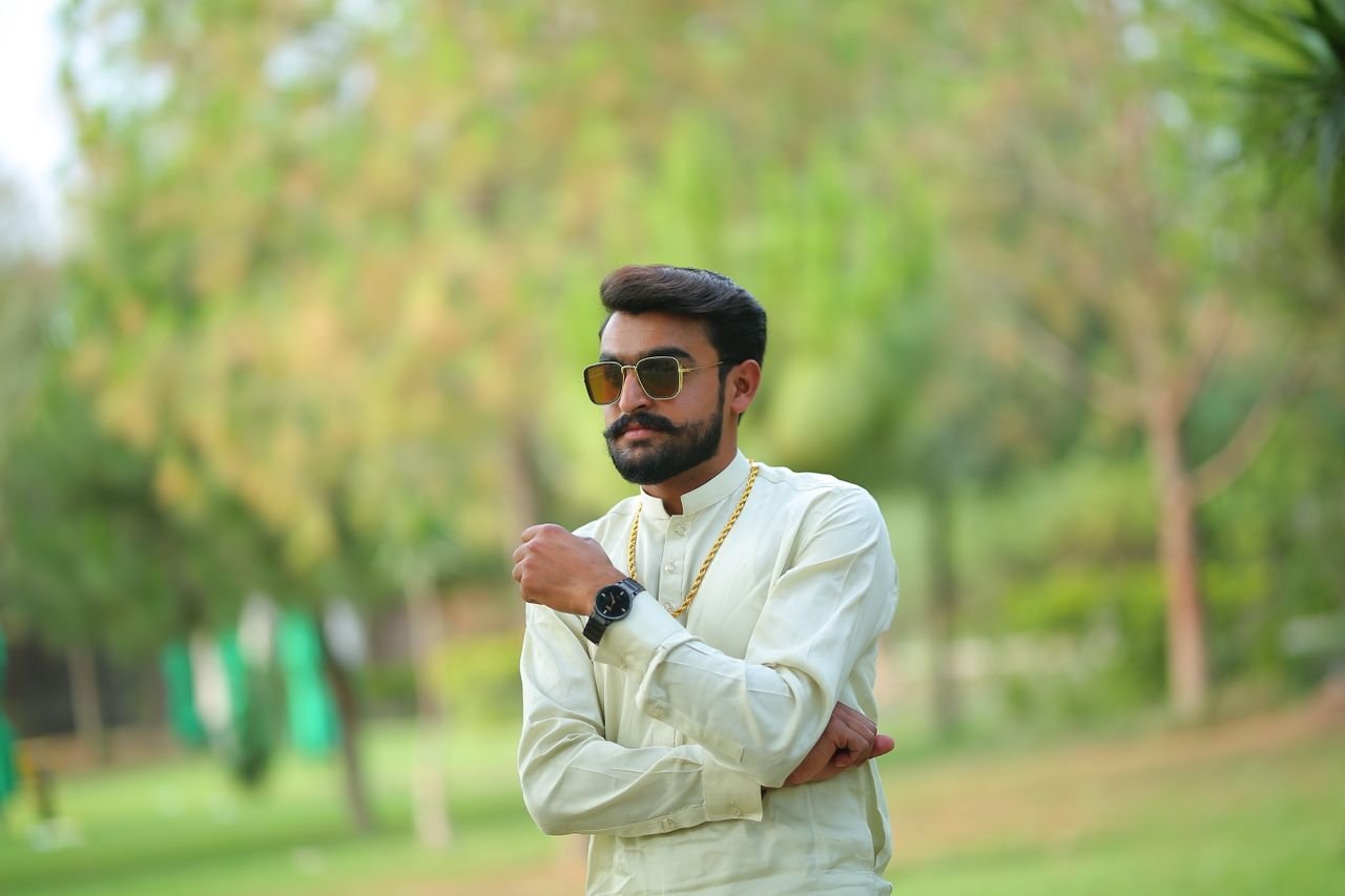 A confident South Asian man poses in traditional attire outdoors in a lush park setting.